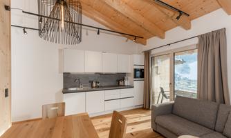 A modern kitchen with a wooden ceiling and a cozy living area. Large windows offer a view of the landscape.
