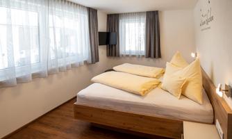 A modern bedroom with a double bed and large windows. The walls are light, and the floor is made of wood.