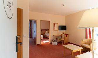 A bright hotel room with a cozy seating area and a table. In the background, another bedroom and a television are visible.