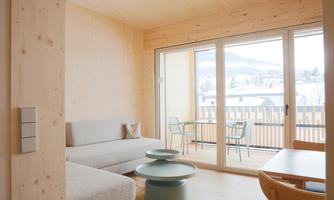 A modern living room with wooden paneling and large windows. The room has a comfortable sofa, a table, and a balcony with a view of the landscape.