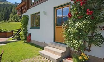 A cozy house entrance with a yellow door and colorful flowers. In the background, green meadows and wooded mountains can be seen.