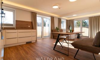 A modern and bright living room with wooden flooring and large windows. The room is stylishly furnished with a dining table and a comfortable sofa.