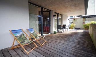 A modern terrace with two lounge chairs and a small table. In the background, large windows and a green area are visible.