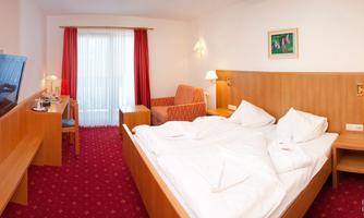 A cozy hotel room with a large bed, a television, and an armchair. The windows let in plenty of light and offer a view of the outdoors.