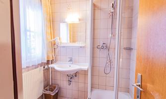 A small bathroom with a shower, a sink, and a mirror. The walls are light-tiled and a window lets in daylight.