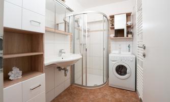 A modern bathroom with a shower and a washbasin. The washing machine is next to a shelf with white and wood-colored design.