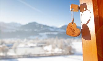 A wooden heart-shaped keychain is hanging on a door. In the background, snow-covered mountains and a blue sky are visible.