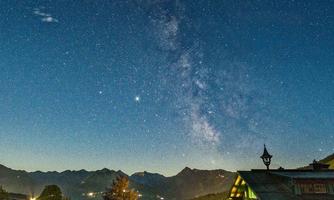 A clear night sky full of stars and the Milky Way above a mountainous landscape. In the foreground, a typical wooden house is visible.