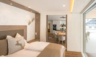 A modern bedroom with a large bed and wooden decor. In the background, an open kitchen is visible.