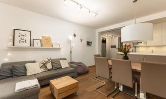 A modern living room with a gray couch, a wooden table, and stylish chairs. The walls are light and adorned with decorative elements.