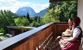 Two women in traditional clothing sit on a balcony and enjoy the view of the mountains. In front of them, a green landscape stretches under a blue sky.