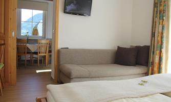 A cozy living room with a gray couch and a TV on the wall. In the background, a dining area and windows with a view outside can be seen.