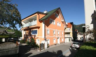 An orange building with several windows and a balcony. It is located on a quiet street, surrounded by trees and other buildings.