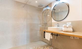 A modern bathroom with a glass shower and a round mirror. Clean towels are ready on the wooden shelf.