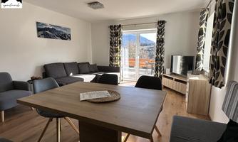 A bright and modern living room with a large window front and mountain view. The decor includes a gray sofa, a wooden table, and a television.