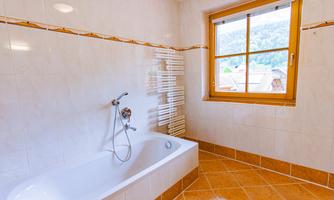 A modern bathroom with a bathtub and a window that offers a view of the mountains. The walls are lightly tiled, and the floor has brown tiles.