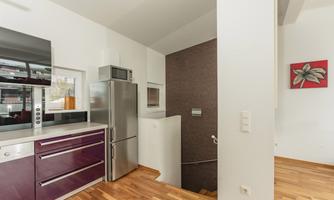 Modern kitchen with a refrigerator, a microwave, and wooden flooring. The walls are painted in neutral colors and there is a minimalist staircase.