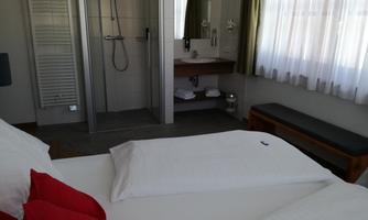 A modern hotel room with two beds and red cushions. In the background, there is a bathroom with a shower and a washbasin.