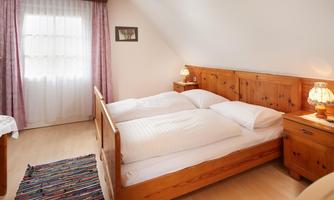 A cozy bedroom with a wooden double bed and two nightstands. The windows are adorned with curtains and the floor is covered with a colorful rug.