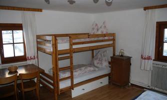 A cozy bedroom with a wooden bunk bed. There is a table and chairs by the window, and the walls are painted in light colors.