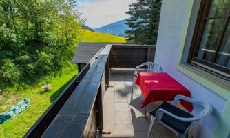 A balcony with a red table and two chairs. In the background, trees and hills can be seen.