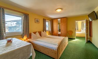 A cozy room with two beds, wooden furniture, and a green carpet. The window offers a view of the surrounding landscape.