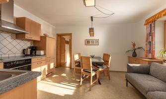 A bright kitchen with a dining area and a gray sofa. The room has wooden cabinets and large windows that provide plenty of light.