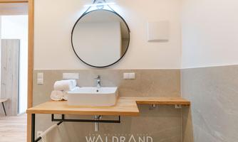 A modern bathroom with a round mirror and a white sink. The walls are in neutral colors, and a wooden shelf complements the stylish design.