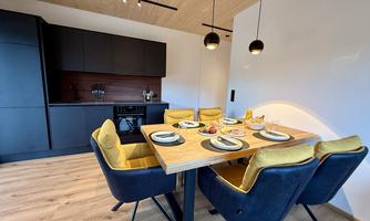 A modern kitchen with a dining table and comfortable chairs. The walls are light and the furniture is in neutral tones.