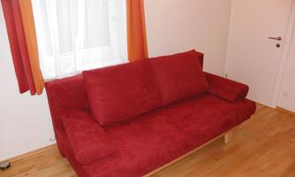 A red sofa with wooden legs stands in a bright room. Daylight falls through the window and there are colored curtains.