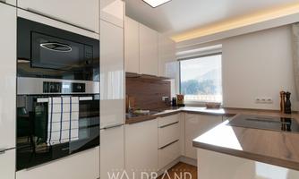 A modern kitchen with white high-gloss cabinets and a wooden countertop.  
Plenty of daylight comes in through the window, creating a bright atmosphere.