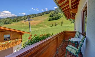 An idyllic view from a balcony overlooking green meadows and gentle hills. In the background, trees and a ski lift can be seen.