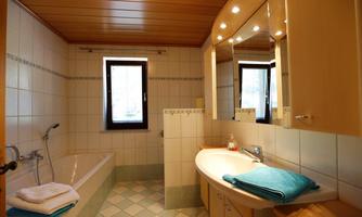A bright bathroom with a bathtub, sink, and large mirrors. The walls are tiled, and the wooden ceiling provides a cozy atmosphere.