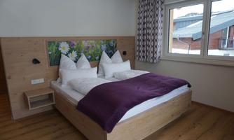 A modern bedroom with a double bed and white pillows. The room has wooden walls, a purple throw, and a large window looking outside.
