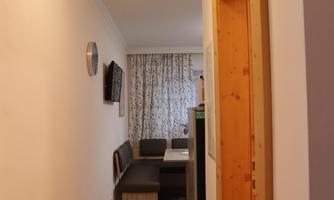 A bright hall with wooden floors and an open door to a living area. A television is mounted on the wall and there is a sofa with a table.