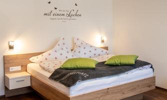 A modern bedroom with a large double bed and a wooden dresser. The walls are simple and there is pleasant lighting.