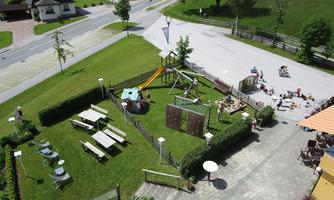 A playground with slides and swings on a green meadow. In the background, there are wooden tables and chairs as well as some people visible.