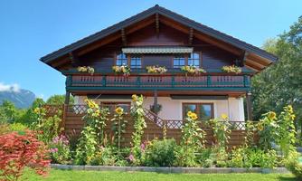 A beautiful, rustic house with a balcony and blooming plants in the garden. It is surrounded by a lush, green environment.