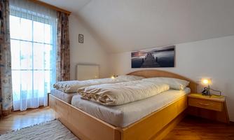 A cozy bedroom with a double bed and large windows. The decoration is simple and the floor is made of wood.