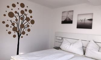 A bright bedroom with a modern bed and two white pillows. On the wall, there is a decorative tree and two black-and-white pictures.