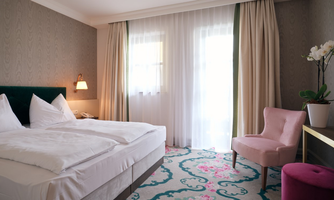A stylish hotel room with a large bed and a pink armchair. The room has bright curtains and a colorful carpet.