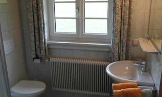 A bright bathroom with a shower, sink, and toilet. Windows with curtains provide daylight.