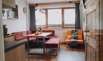 A cozy apartment with a living area that includes a dining corner and a comfortable armchair. Large windows provide plenty of daylight.