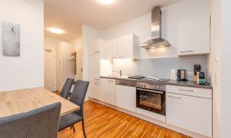 A modern kitchen with white furniture and wooden flooring. There is a dining table and chairs as well as a kitchenette with appliances.