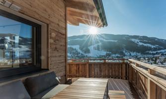 A cozy balcony terrace with a wooden railing andsun rays shining over snow-covered mountains. In the foreground, there is a wooden log table and a comfortable seating area.