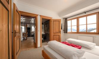 A cozy bedroom with wooden details and a large window. In the background, another room is visible, radiating a rustic atmosphere.