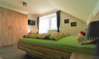 A cozy bedroom with a wooden bed and colorful bedding. The room has large windows and a television on the wall.