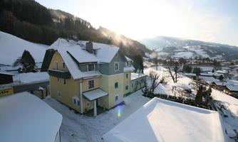 A cozy house in a wintry landscape, surrounded by snow-covered hills. The sun is shining, lending a warm atmosphere to the scene.