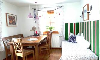 A cozy living room with a wooden table and chairs. The walls are decorated and a window with plants brings light in.
