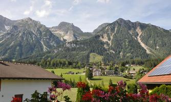 A picturesque mountain landscape with high peaks and lush forests. In the foreground, colorful flowers bloom and a typical alpine building is visible.